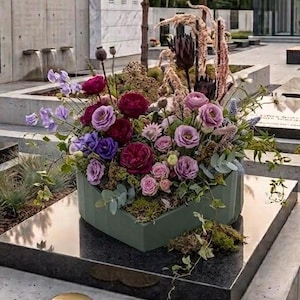 May include: A floral arrangement in a sage green, faceted, heart-shaped container. The bouquet features deep burgundy and light purple roses, lavender sweet peas, dried grasses, and eucalyptus leaves, displayed on a polished black stone surface.