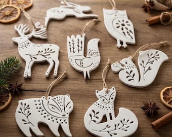 May include: A collection of seven white ceramic animal ornaments featuring intricate etched floral and geometric patterns. The set includes a deer, rabbit, owl, bird, squirrel, bear, and fox, each suspended by a thin tan twine string.