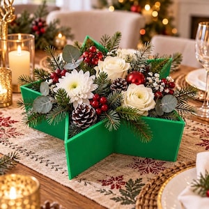 Könnte beinhalten: Ein sternförmiger grüner Behälter mit einem weihnachtlichen Blumenarrangement aus weißen Rosen, weißen Chrysanthemen, roten Beeren, Tannenzapfen, Tannenzweigen, Eukalyptusblättern und einer einzelnen glänzenden roten Weihnachtskugel.