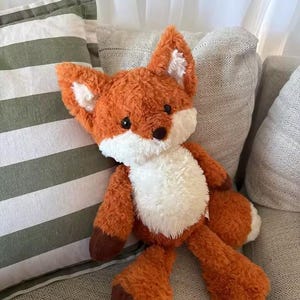 May include: A plush fox toy with orange and white fur, featuring dark brown paws, black eyes, and a black nose. The stuffed animal has pointed ears with white inner fur and a white chest patch, designed with a soft, textured, shaggy fabric finish.