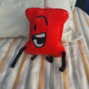 May include: A plush red pushpin character with a smiling face, black legs, and a white and gray mouth. The pushpin has a white, cone-shaped top. The plush is on a striped bedspread.