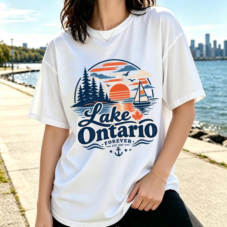 May include: A white short-sleeve t-shirt featuring a graphic print of a sunset over Lake Ontario. The design includes a sailboat, pine trees, a maple leaf, and an anchor with the text Lake Ontario FOREVER EST. 1867 in blue and orange.