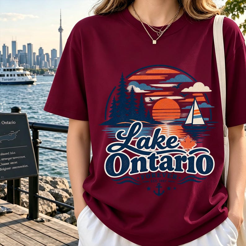 May include: A maroon short-sleeved t-shirt featuring a graphic print of a sunset over Lake Ontario with pine trees, a sailboat, and a maple leaf. The text reads Lake Ontario Forever Est 1783 with an anchor icon below the lettering.