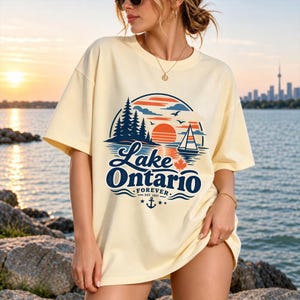 May include: A cream-colored short-sleeve t-shirt featuring a graphic of a sunset over a lake with pine trees, a sailboat, and a maple leaf. The text reads Lake Ontario FOREVER EST. 1867, accompanied by an anchor icon and small stars.