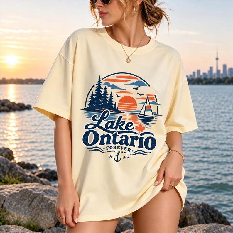 May include: A cream-colored short-sleeve t-shirt featuring a graphic of a sunset over a lake with pine trees, a sailboat, and a maple leaf. The text reads Lake Ontario FOREVER EST. 1867, accompanied by an anchor icon and small stars.