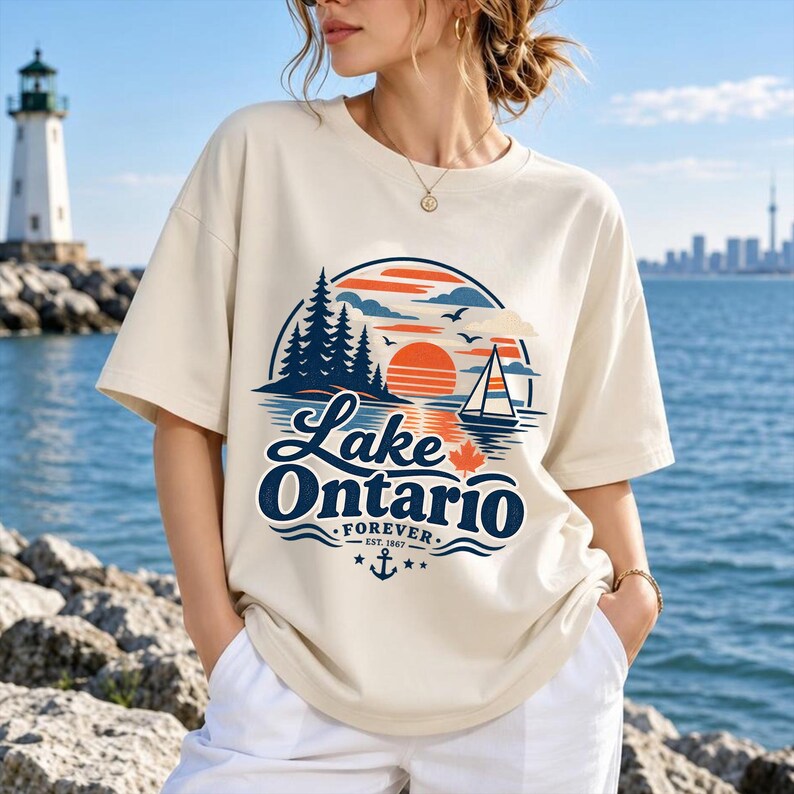 May include: A cream-colored short-sleeve t-shirt featuring a graphic of a sunset over water, a sailboat, pine trees, and a maple leaf. The text reads Lake Ontario Forever Est. 1867 with an anchor icon and stars. The design uses orange, blue, and white.