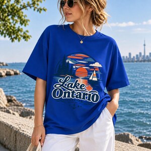 May include: A royal blue short-sleeve t-shirt featuring a graphic print of a sunset over water with a sailboat, pine trees, and a small maple leaf. The text reads Lake Ontario Forever with an anchor icon centered below the lettering.