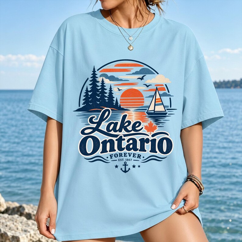 May include: A light blue t-shirt featuring a graphic print of a sunset over Lake Ontario. The design includes a sailboat, pine trees, a maple leaf, and the text "Lake Ontario FOREVER EST. 1867" with an anchor icon centered below the lettering.