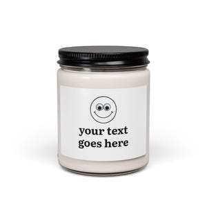 Op de afbeelding: Een doorzichtige glazen kaarsenpot met een zwarte metalen deksel, gevuld met gebroken witte was. De voorzijde heeft een wit rechthoekig etiket met een zwart smiley-icoon boven de dikgedrukte tekst, your text goes here.