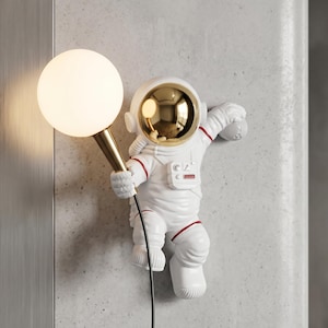 Könnte beinhalten: Eine weiße Wandlampe in Astronautenform mit goldenen Akzenten und einem runden, leuchtenden Licht. Der Astronaut hält das Licht an einem goldenen Stiel, mit roten Details am Anzug. Ein schwarzes Kabel geht von der Lampe ab.