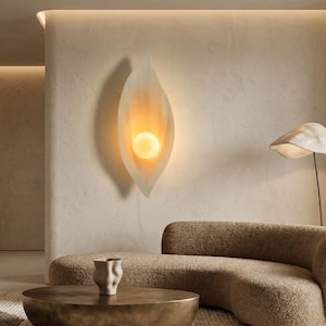 May include: A contemporary wall light with a leaf-inspired form. The sconce has a frosted glass outer layer and a warm, radiant light. It's mounted on a textured, neutral-toned wall, enhancing the room's aesthetic with a touch of sophistication.