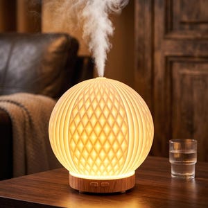 May include: A spherical, illuminated essential oil diffuser with a diamond pattern and a wooden base. The diffuser emits a stream of vapour. A glass of water sits to the right. The background features a leather couch and wooden door.