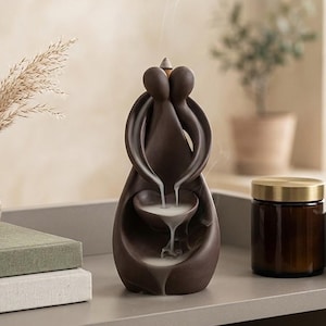 May include: A brown ceramic incense waterfall with a couple embracing design. The incense smoke flows down into a small bowl. A small vase with dried grass and a jar with a gold lid are also on the shelf.