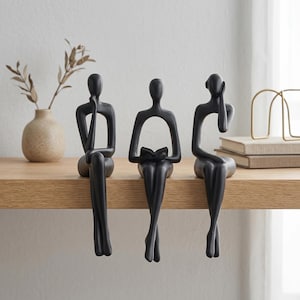 Pode incluir: Três esculturas abstratas pretas de figuras sentadas, cada uma em uma pose diferente: uma com a mão no queixo, outra lendo um livro e outra com a mão na cabeça. Elas são exibidas em uma prateleira de madeira.