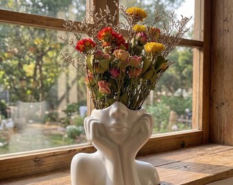 May include: A white ceramic vase shaped like a human head resting on hands, filled with a bouquet of dried flowers in shades of red, yellow, and pink. The vase features a smooth, glossy finish and is positioned in front of a wooden window frame.