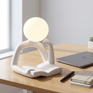 May include: A white desk lamp shaped like a person reading a book, with a round, glowing light bulb for a head. The lamp is on a wooden desk, with a laptop, pen, and notebook nearby.