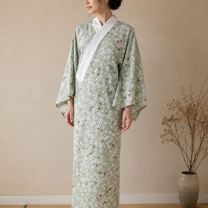 May include: A sage green yukata featuring a repeating pattern of small white flowers with dark centers. The garment has wide, flowing sleeves and a solid white collar that wraps across the front, extending down to the floor in a straight silhouette.
