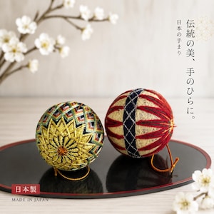 以下が含まれることがあります： 複雑な刺繍が施された2つの手まり。1つは黒地に黄色の星模様と緑と白の幾何学的なアクセントが特徴です。もう1つは赤とクリーム色のストライプに、紺色のクロスステッチの帯が施されています。伝統の美、手のひらに。日本製。