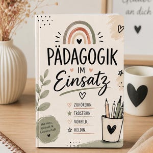 Könnte beinhalten: Ein Notizbuch mit cremefarbenem Einband, einer Regenbogen-Illustration, dem schwarzen Text PÄDAGOGIK IM Einsatz und einer Liste mit den Begriffen ZUHÖRERIN, TRÖSTERIN, VORBILD und HELDIN. Es enthält eine Stiftebecher-Grafik und ein rundes Abzeichen.