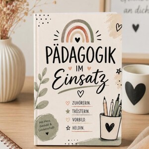 Könnte beinhalten: Ein beigefarbenes Notizbuch mit einer Regenbogen-Illustration und dem Text PÄDAGOGIK IM Einsatz. Darunter stehen die Wörter ZUHÖRERIN, TRÖSTERIN, VORBILD und HELDIN sowie eine Zeichnung eines Stiftehalters. Ein runder Aufkleber zeigt Mit Herz, Geduld & Leidenschaft.