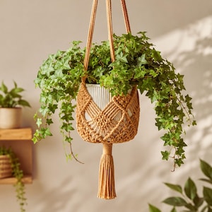 May include: A mustard yellow macrame plant hanger featuring a diamond-patterned knot design and a long tassel at the bottom. The hanger holds a white, vertically ribbed cylindrical pot containing a lush, trailing green ivy plant.