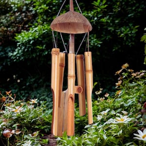 Op de afbeelding: Een bamboe windgong met een natuurlijke afwerking hangt aan een ketting. De windgong heeft een kokosnoot schaal bovenkant en verschillende bamboe buizen van verschillende lengtes. De achtergrond toont weelderig groen gebladerte en witte bloemen.