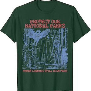 Peut inclure: Un t-shirt vert foncé à manches courtes avec une impression graphique bleue représentant une scène de forêt avec une chauve-souris, un sasquatch, des fantômes, une créature ressemblant à un cerf et le texte PROTECT OUR NATIONAL PARKS WHERE LEGENDS STILL ROAM FREE en rouge.
