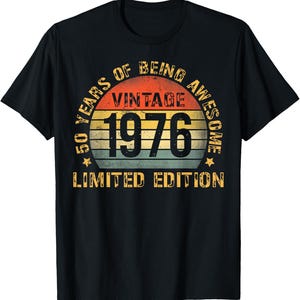 Può includere: Una t-shirt nera a maniche corte con una grafica circolare su uno sfondo a strisce arancioni, gialle e blu che ricordano un tramonto. Il testo recita 50 YEARS OF BEING AWESOME, VINTAGE 1976 e LIMITED EDITION in un carattere giallo.
