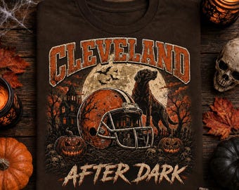 May include: A dark brown t-shirt featuring a graphic of a football helmet, a dog, a haunted house, and pumpkins under a full moon. The text CLEVELAND is arched above in orange, and the words AFTER DARK are printed in a distressed font below.
