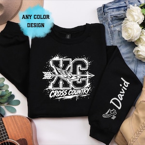 May include: A black long-sleeve sweatshirt featuring a white graphic with the text XC CROSS COUNTRY, an arrow, and a winged shoe. The sleeve displays the name David in white script above a small shoe icon. A blue cloud graphic reads ANY COLOR DESIGN.