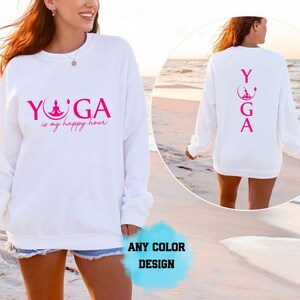 Puede incluir: Una sudadera blanca de manga larga con texto y gráfico en rosa intenso. El frente muestra la palabra YOGA con una figura en posición de loto dentro de la O, seguida del texto is my happy hour. La parte posterior muestra la palabra YOGA vertical.