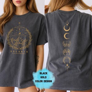 Pode incluir: Camiseta cinza escuro com gr&aacute;ficos dourados na frente e nas costas. O design frontal apresenta uma cena de montanha dentro de uma coroa, com o texto "VELARIS - CITY OF STARLIGHT". A parte de tr&aacute;s tem um design vertical de luas e estrelas.