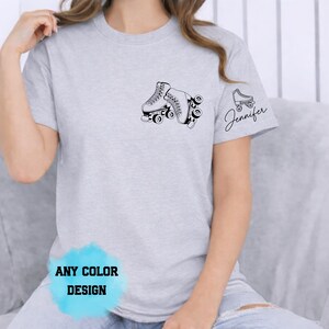 May include: A light gray t-shirt featuring a black roller skate graphic on the chest and a smaller skate with the name "Jennifer" on the sleeve. The shirt has a crew neck and short sleeves. The text "ANY COLOR DESIGN" is at the bottom.