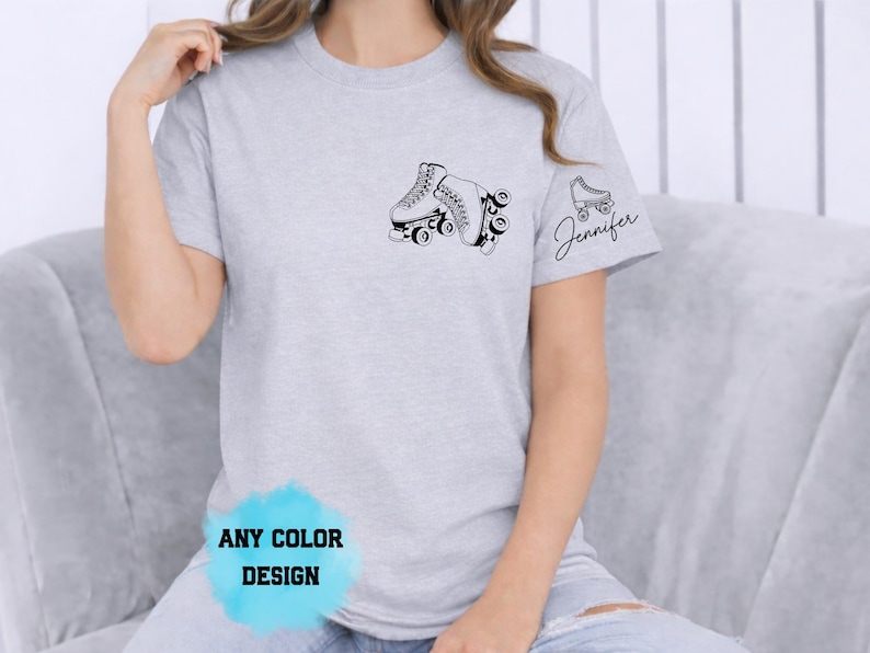 May include: A light gray t-shirt featuring a black roller skate graphic on the chest and a smaller skate with the name "Jennifer" on the sleeve. The shirt has a crew neck and short sleeves. The text "ANY COLOR DESIGN" is at the bottom.