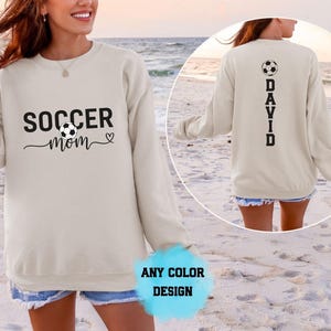 Puede incluir: Una sudadera beige de manga larga con el texto negro SOCCER mom, un gráfico de balón de fútbol y un corazón. La parte posterior de la sudadera muestra el nombre DAVID en letras negras verticales con un gráfico de balón de fútbol encima.