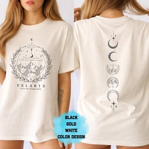 Pode incluir: Camiseta creme com um gr&aacute;fico frontal com montanhas, texto e um design de coroa de louros. A parte de tr&aacute;s da camiseta exibe um gr&aacute;fico vertical de fases da lua. O texto na frente diz "VELARIS - CITY OF STARLIGHT".