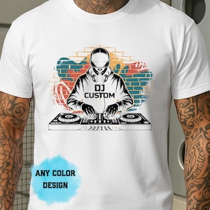 May include: White t-shirt with a black and white graphic of a DJ at a mixing console. The design includes the text "DJ CUSTOM" and a colourful graffiti-style background. The shirt also has the text "ANY COLOUR DESIGN".