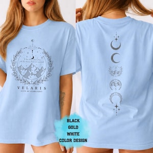 Pode incluir: Camiseta azul clara com um gr&aacute;fico frontal com montanhas, uma lua crescente e o texto "VELARIS - CITY OF STARLIGHT". A parte de tr&aacute;s da camisa exibe um design vertical de luas crescentes e estrelas. Dispon&iacute;vel em preto, dourado e branco.