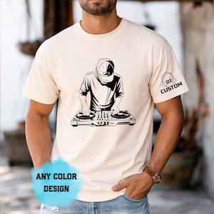 Op de afbeelding: Een crèmekleurig T-shirt met korte mouwen en een zwarte afbeelding van een DJ met een pet die een draaitafelconsole gebruikt. De linkermouw toont de tekst DJ CUSTOM met een koptelefoonpictogram. Een blauwe grafische overlay leest ANY COLOR DESIGN.