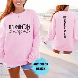 Puede incluir: Una sudadera de manga larga de color rosa claro con texto negro en la parte delantera que dice BADMINTON MOM junto a un corazón y un volante. La parte trasera de la sudadera muestra el nombre OLIVIA en vertical con un icono de volante.
