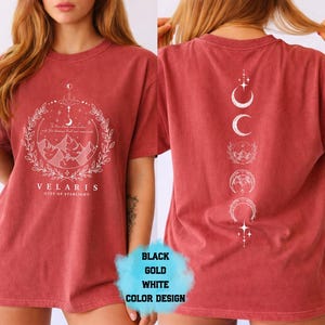 Pode incluir: Camiseta vermelho-alaranjada com um gr&aacute;fico branco na frente com montanhas, uma lua crescente e o texto "VELARIS - CITY OF STARLIGHT". A parte de tr&aacute;s da camisa tem um desenho branco de fases da lua e estrelas.