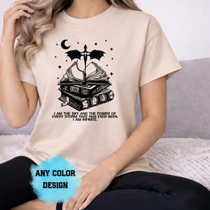 May include: Beige t-shirt with a black graphic design featuring a stack of books, a sword, a crescent moon, and stars. The text "I AM THE SKY AND THE POWER OF EVERY STORM THAT HAS EVER BEEN. I AM INFINITE." is below the graphic. The words "ANY COLOR DESIGN" are at the bottom.