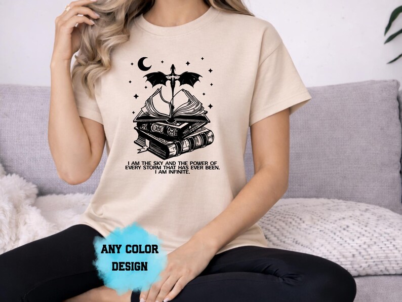 May include: Beige t-shirt with a black graphic design featuring a stack of books, a sword, a crescent moon, and stars. The text "I AM THE SKY AND THE POWER OF EVERY STORM THAT HAS EVER BEEN. I AM INFINITE." is below the graphic. The words "ANY COLOR DESIGN" are at the bottom.