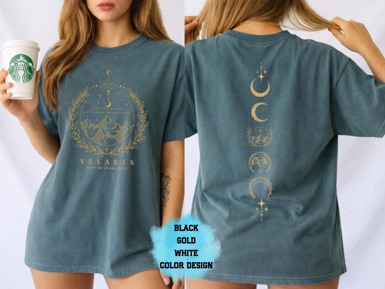 Pode incluir: Camiseta azul-petr&oacute;leo com um gr&aacute;fico dourado na frente com montanhas, uma lua crescente e a palavra "VELARIS". A parte de tr&aacute;s da camisa exibe um design vertical de luas crescentes e estrelas douradas. A camiseta &eacute; de estilo casual.