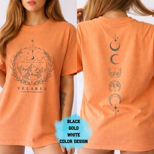 Pode incluir: Camiseta laranja com um gr&aacute;fico frontal com montanhas, uma lua crescente e o texto "VELARIS - CITY OF STARLIGHT". A parte de tr&aacute;s da camisa exibe um design vertical de fases lunares. A camisa tem um ajuste folgado.