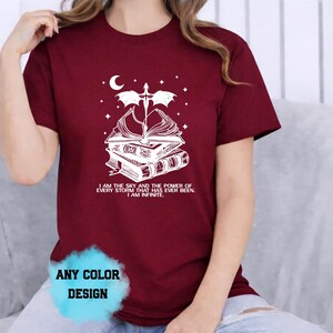 May include: Burgundy t-shirt with a white graphic of books, a sword, a crescent moon, and stars. The text on the shirt reads "I AM THE SKY AND THE POWER OF EVERY STORM THAT HAS EVER BEEN. I AM INFINITE."