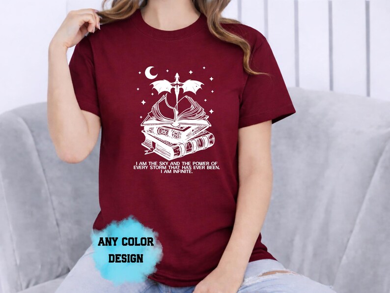 May include: Burgundy t-shirt with a white graphic of books, a sword, a crescent moon, and stars. The text on the shirt reads "I AM THE SKY AND THE POWER OF EVERY STORM THAT HAS EVER BEEN. I AM INFINITE."