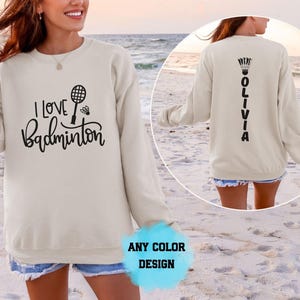 May include: A beige long-sleeve sweatshirt featuring black text on the front that reads I LOVE BADMINTON with a racket and shuttlecock graphic. The back of the sweatshirt displays the name OLIVIA vertically with a shuttlecock icon above the text.
