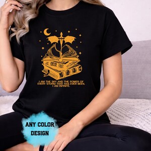 May include: Black t-shirt with a gold graphic featuring a stack of books, a sword, a crescent moon, and stars. The text reads "I AM THE SKY AND THE POWER OF EVERY STORM THAT HAS EVER BEEN. I AM INFINITE." The shirt design is available in any color.