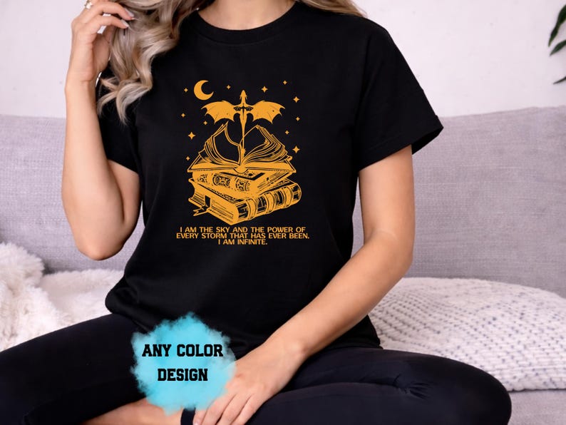 May include: Black t-shirt with a gold graphic featuring a stack of books, a sword, a crescent moon, and stars. The text reads "I AM THE SKY AND THE POWER OF EVERY STORM THAT HAS EVER BEEN. I AM INFINITE." The shirt design is available in any color.